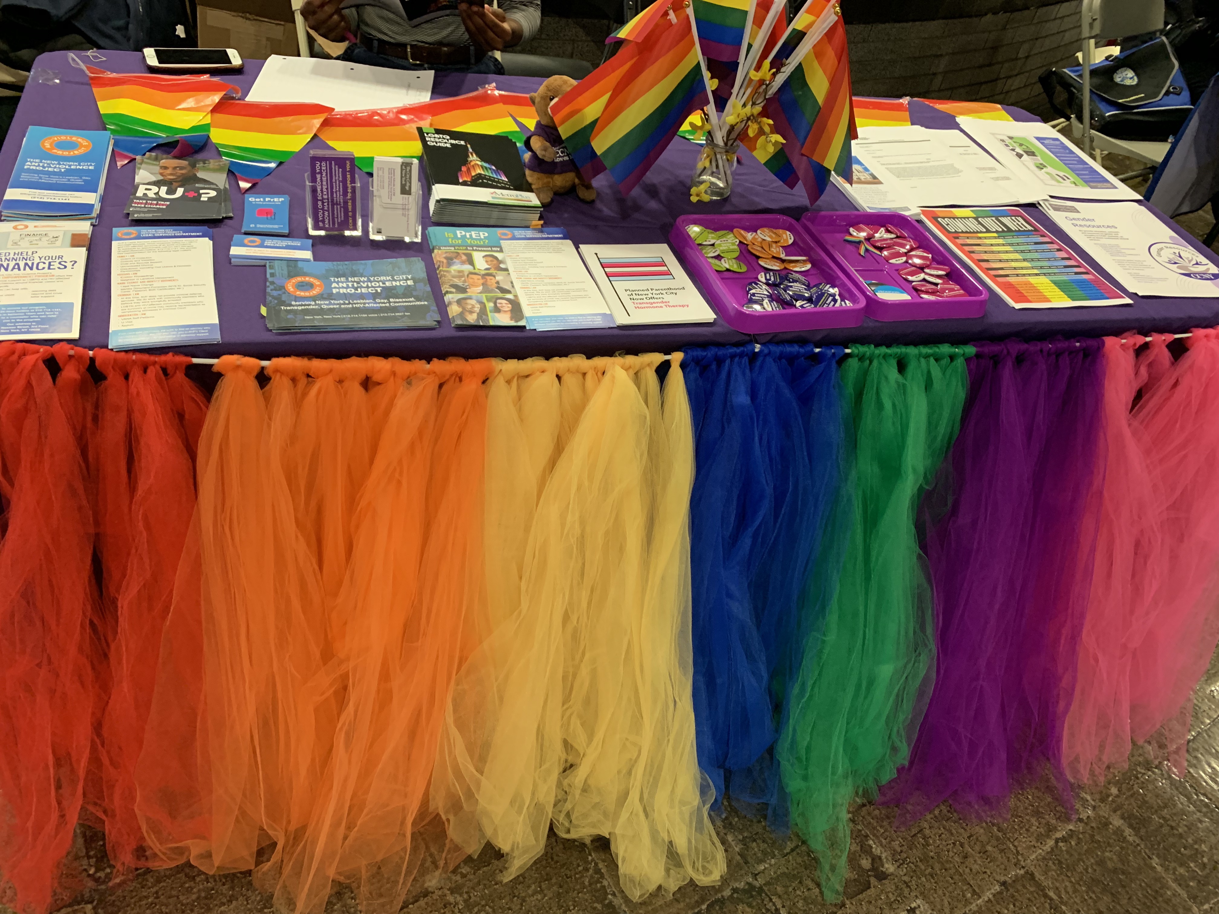 This image showcases a table display from CCNYs 'Coming out Week' event. The table is full of the rainbow colors of the pride flag, and includes pamphlets and fliers of different resources.