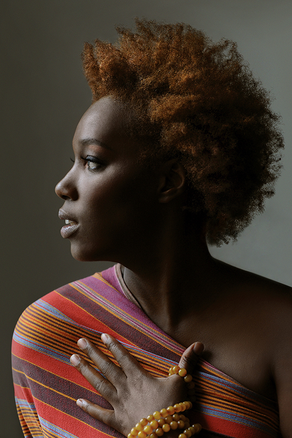 Imani Uzuri A Black womxn faces left while the light shines on their copper curls, multi-colored dress, and orange stone jewelry. Their left hand sits open on their chest and they appear to be looking in the distance.