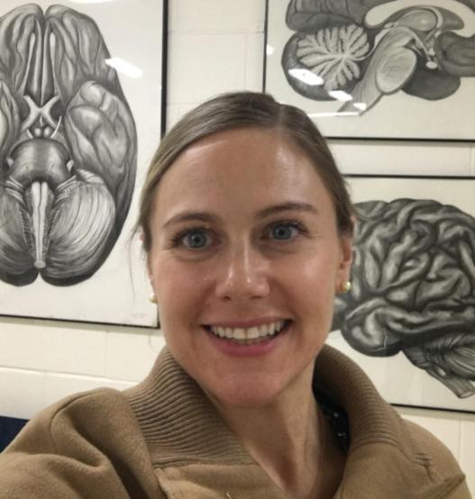 Self-portrait of smiling professor in front of large medical drawings of brains, taken in the hallway outside of the Biology Department.