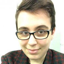 A profile photo of a young white woman with brown hair and glasses. She is smiling and wearing a red and green blazer over a black T-shirt.