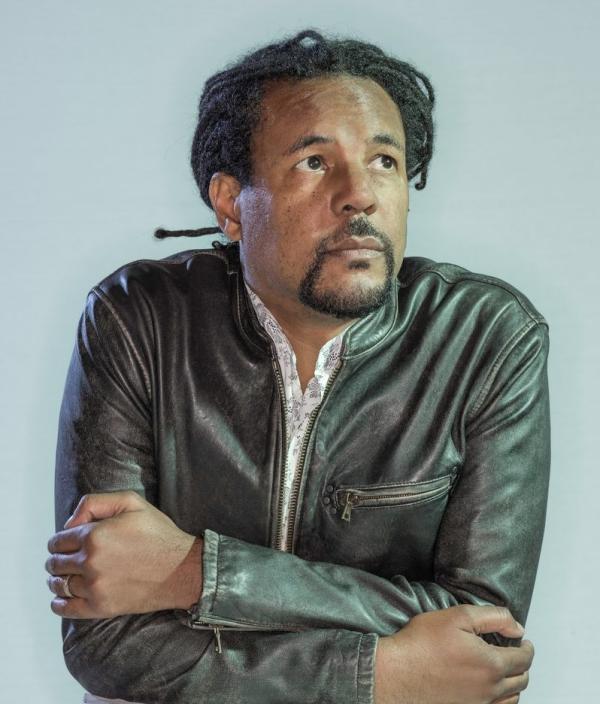 Colson Whitehead looking of to the side in a leather jacket with crossed arms.