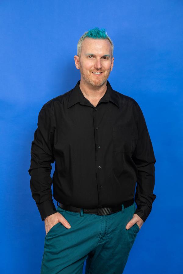 Bryan Stanton, average height, slightly chubby, white person, with blonde hair and blue streaks standing wearing teal pants and a black long sleeve shirt. Their hands at their sides.