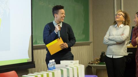 Three people are standing on stage looking at a screen - all three are smiling and joking around 