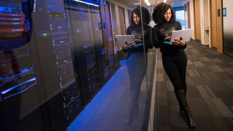 Woman leaning against a glass wall and holding a computer. behind the class wall is a data bank that is glowing a somber blue color. 