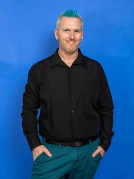Bryan Stanton, average height, slightly chubby, white person, with blonde hair and blue streaks standing wearing teal pants and a black long sleeve shirt. Their hands at their sides.